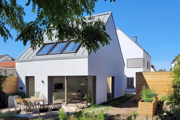 Zwei weiß verputzte Gebäudekörper mit Satteldächern, großen Glasflächen und Gartenbereich mit Terrasse