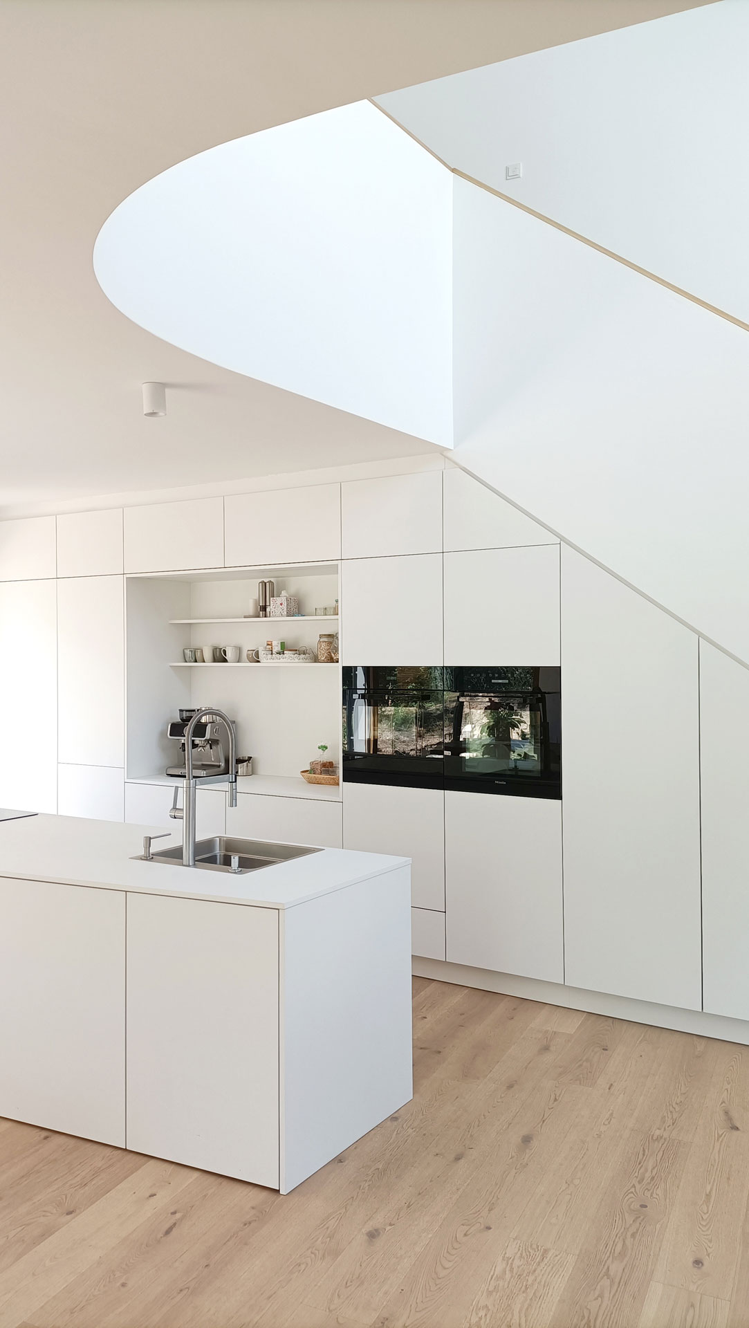 Moderne weiße Einbauküche mit Kücheninsel, hellem Eichenholzfußboden und skulpturaler Treppenöffnung im Deckenbereich.