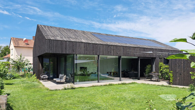 Gartenseite von Haus ANA mit Terrassenlandschaft, Panoramafenster, PV-Anlage und Holzfassade auf der Dachschräge.